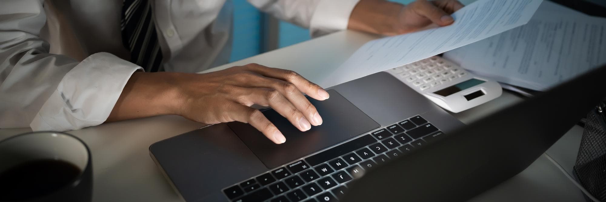 An operator at a desk comparing two printed product brochures and a laptop dashboard, daylight from the side